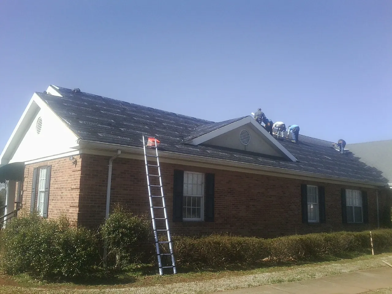 Crew tearing shingles off a brick ranch roof with a ladder set — Metal Roof Repair work by our crew in Le Mars