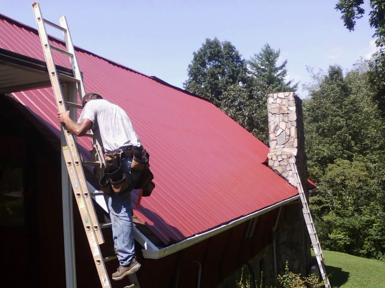 Roofer on a ladder beside a red metal roof and stone chimney — Soffit & Fascia Repair by Roofing Brothers in Le Mars